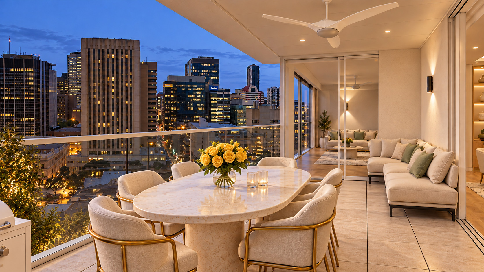 A covered balcony at dusk with a marble dining table, gold-framed chairs and yellow roses, a lounge beside the glass doors and the lit Adelaide city skyline beyond.